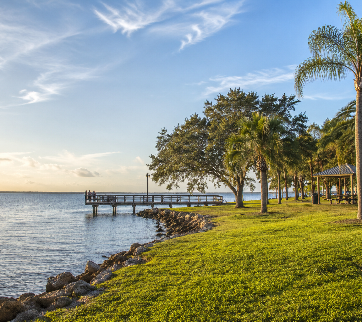 Waterfront park at golden hour with a fishing pier, rock shoreline, palms and a picnic pavilion on open lawn