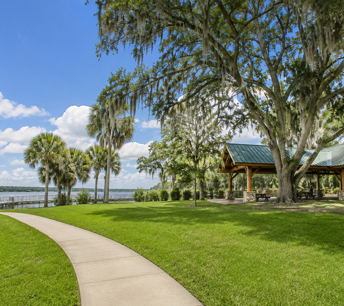 Park beside open water with timber picnic pavilions under live oaks draped in Spanish moss, and a curving path to the shore