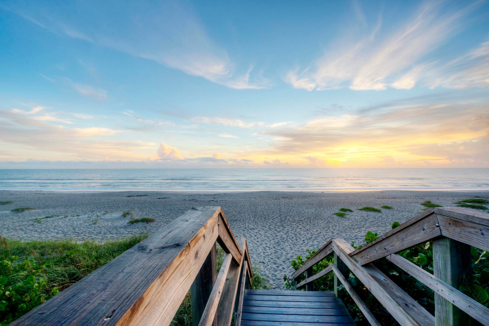 Wooden boardwalk steps down to an empty beach, dune grass on either side and open ocean beyond